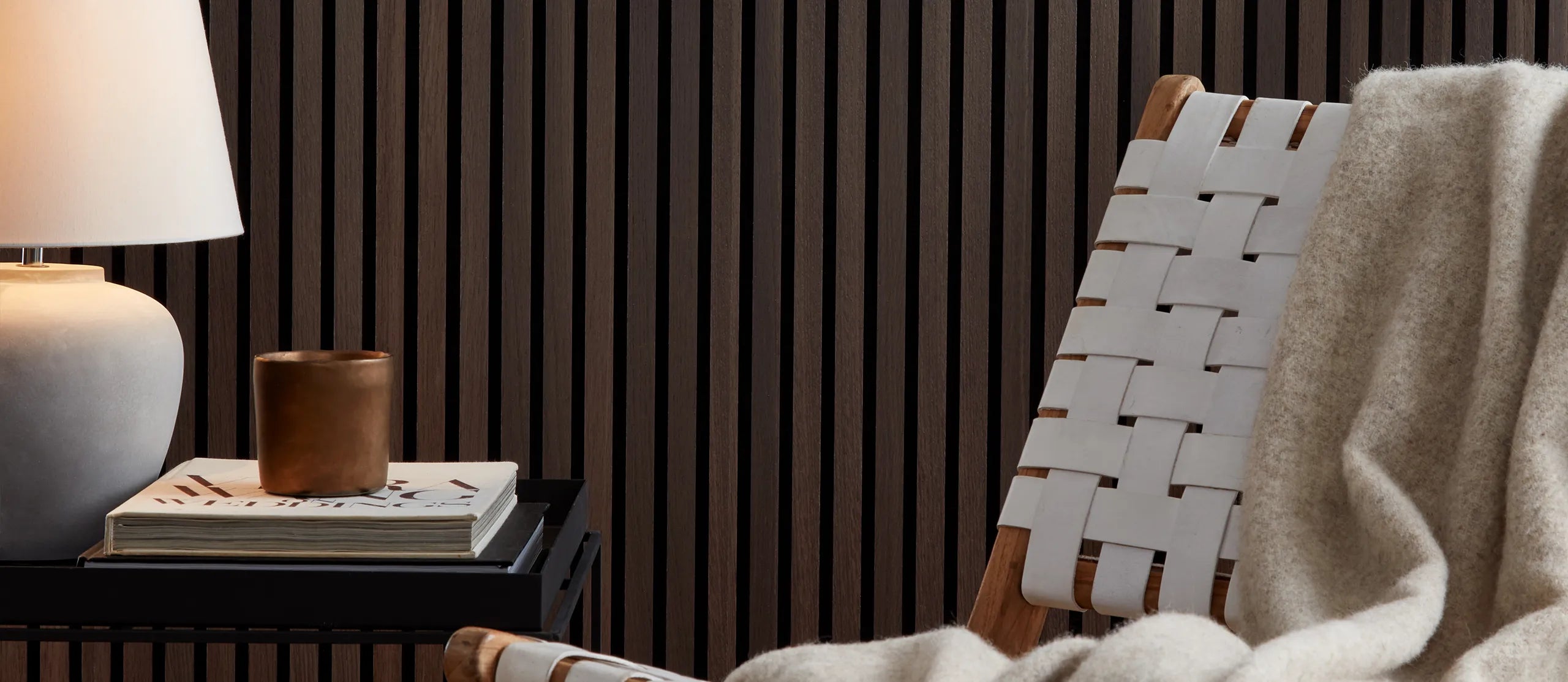 White chair next to small table with lamp, books and and a candle. SlatWall Smoked Oak on the wall in the background